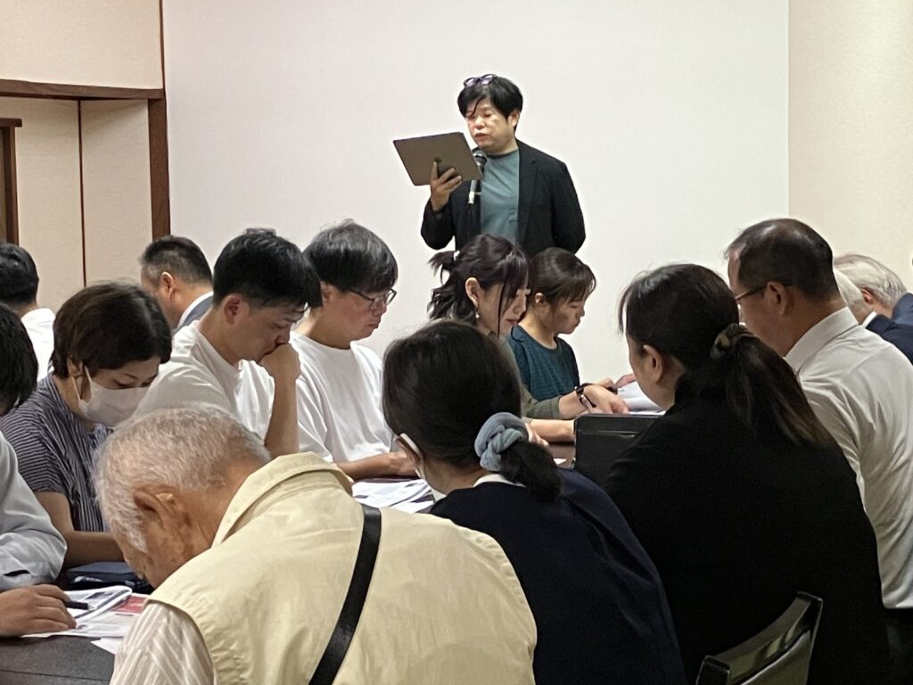 マイクの前で話す当センター代表と、手前で資料を読む参加者の皆さま